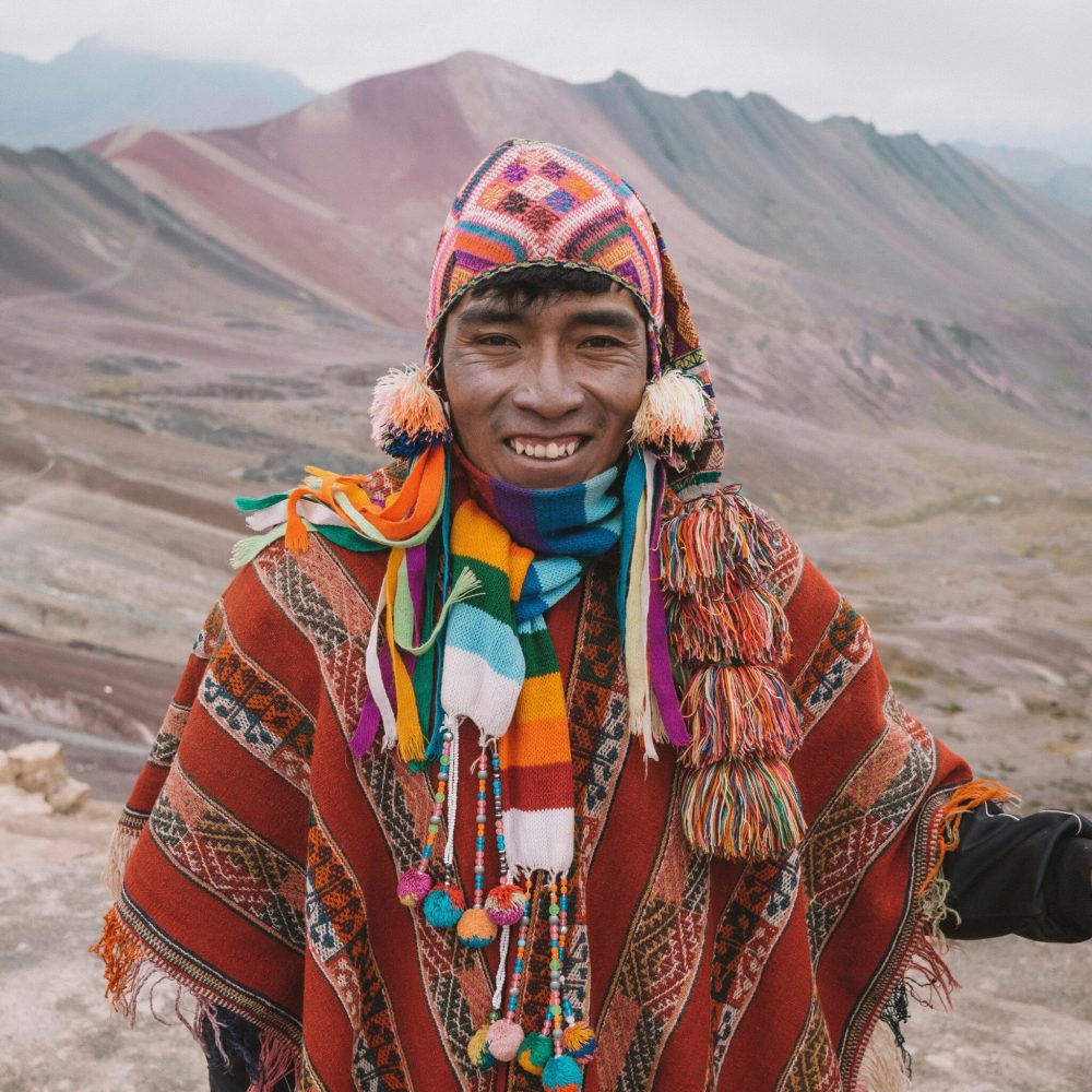 Man in colorful traditional wear in front of Peruvian mountain scenery.